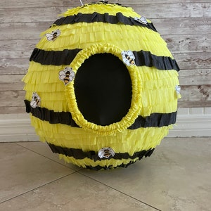 May include: A spherical pi&ntilde;ata designed to resemble a bee. The pi&ntilde;ata is yellow with black stripes and decorative bee stickers. It has a large, circular opening and a white loop for hanging. Perfect for a bee-themed party.