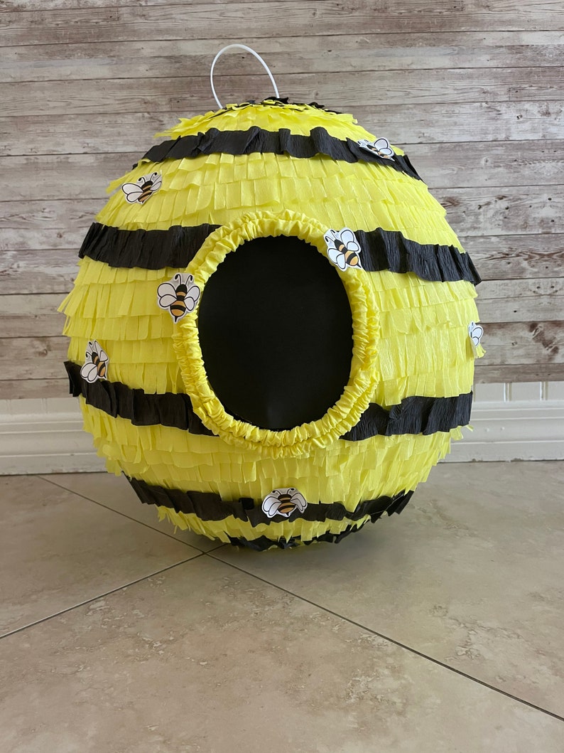May include: A spherical pi&ntilde;ata designed to resemble a bee. The pi&ntilde;ata is yellow with black stripes and decorative bee stickers. It has a large, circular opening and a white loop for hanging. Perfect for a bee-themed party.