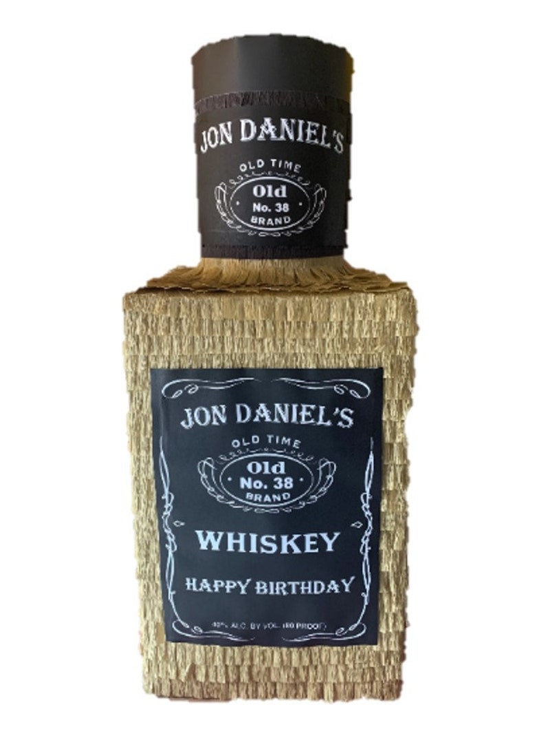 K&ouml;nnte beinhalten: Eine rechteckige Pi&ntilde;ata in Form einer Whiskeyflasche, bedeckt mit goldenem Fransenpapier. Sie hat schwarze Etiketten mit wei&szlig;em Text, auf denen JON DANIEL'S, OLD TIME, Old No. 38 BRAND, WHISKEY und HAPPY BIRTHDAY steht, sowie einen schwarzen zylindrischen Hals.