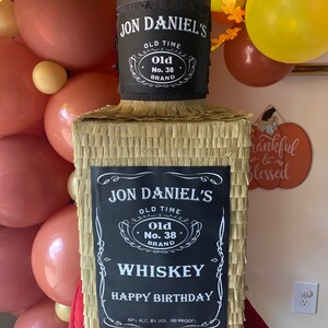 K&ouml;nnte beinhalten: Eine rechteckige Pinata in Form einer Whiskeyflasche, bedeckt mit goldenem Fransenpapier. Sie hat ein schwarzes Etikett mit wei&szlig;em Text: JON DANIEL'S OLD TIME OLD NO. 38 BRAND WHISKEY HAPPY BIRTHDAY 40% ALC. BY VOL. (80 PROOF) und einen schwarzen Flaschenhals.
