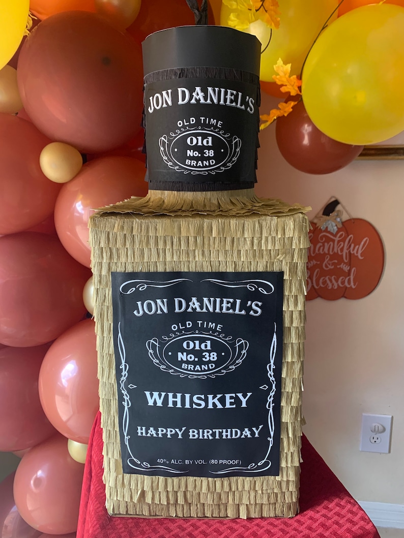 K&ouml;nnte beinhalten: Eine rechteckige Pinata in Form einer Whiskeyflasche, bedeckt mit goldenem Fransenpapier. Sie hat ein schwarzes Etikett mit wei&szlig;em Text: JON DANIEL'S OLD TIME OLD NO. 38 BRAND WHISKEY HAPPY BIRTHDAY 40% ALC. BY VOL. (80 PROOF) und einen schwarzen Flaschenhals.