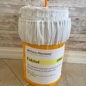 May include: A cylindrical pi&ntilde;ata shaped like a prescription pill bottle with yellow fringe on the sides and white fringe on the top. A label reads Old Farts Pharmacy, Fukitol 100mg, with instructions to take a handful when feeling tired or old.