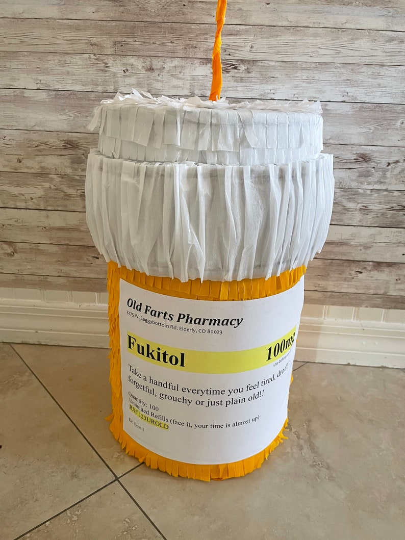 May include: A cylindrical pi&ntilde;ata shaped like a prescription pill bottle with yellow fringe on the sides and white fringe on the top. A label reads Old Farts Pharmacy, Fukitol 100mg, with instructions to take a handful when feeling tired or old.