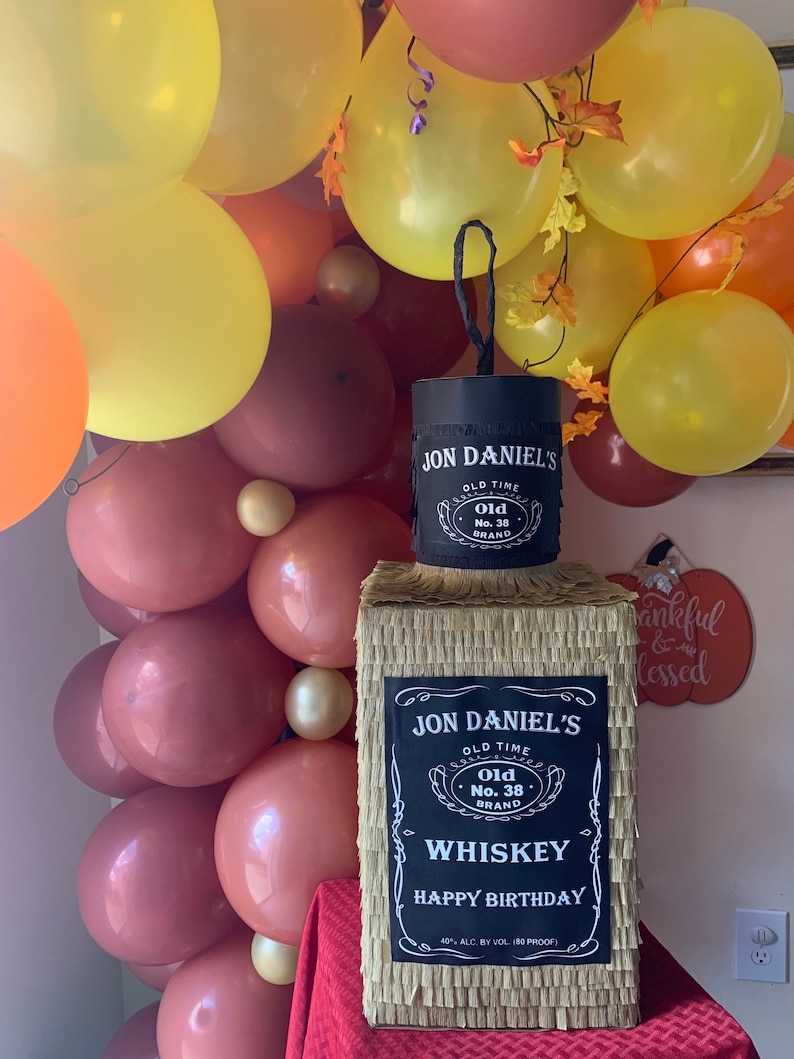 K&ouml;nnte beinhalten: Eine rechteckige Pi&ntilde;ata in Form einer Whiskeyflasche, bedeckt mit beigefarbenem Fransenpapier. Sie hat ein schwarzes Etikett mit wei&szlig;em Text: JON DANIEL'S OLD TIME OLD NO. 38 BRAND WHISKEY HAPPY BIRTHDAY 40% ALC. BY VOL. (80 PROOF).