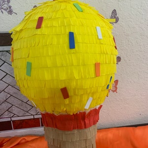 May include: A yellow cupcake-shaped pi&ntilde;ata with colorful confetti accents. The top is made of yellow crepe paper, the base is brown, and the middle is red. A festive party decoration.