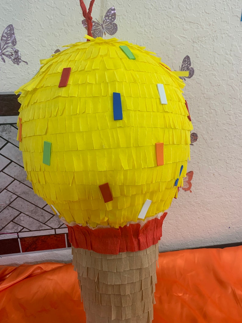 May include: A yellow cupcake-shaped pi&ntilde;ata with colorful confetti accents. The top is made of yellow crepe paper, the base is brown, and the middle is red. A festive party decoration.