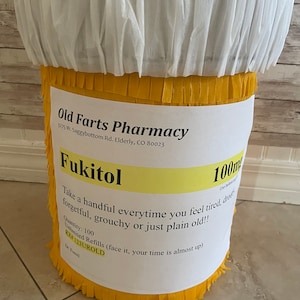 May include: A cylindrical pi&ntilde;ata designed to resemble a prescription pill bottle with a white top and yellow fringed body. A white label features the text Old Farts Pharmacy, Fukitol 100mg, and instructions to take a handful when feeling old.