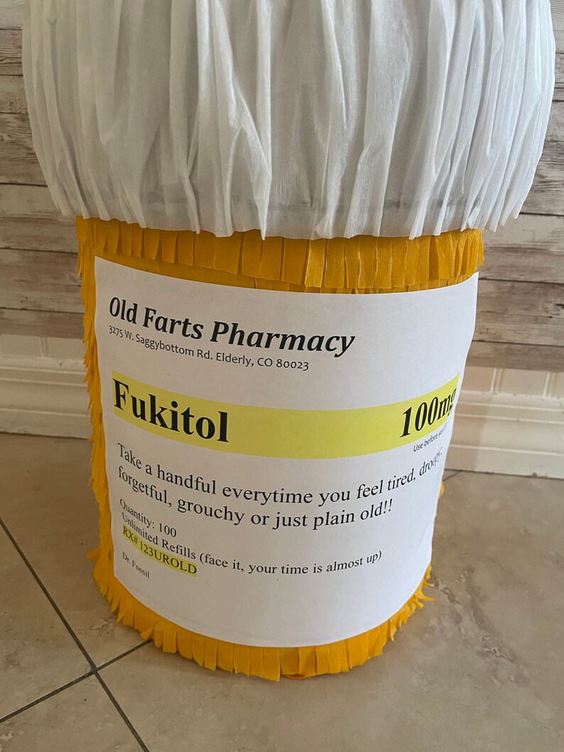 May include: A cylindrical pi&ntilde;ata designed to resemble a prescription pill bottle with a white top and yellow fringed body. A white label features the text Old Farts Pharmacy, Fukitol 100mg, and instructions to take a handful when feeling old.