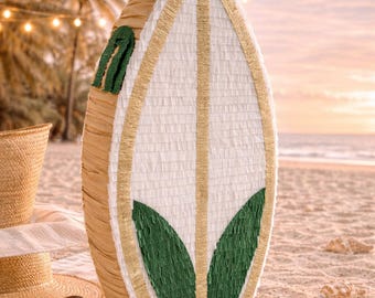 May include: A surfboard-shaped piñata featuring a white center panel with gold trim and two dark green leaf-shaped accents. The sides are covered in tan fringe, and a white hanging loop is attached to the top of the vertical structure.