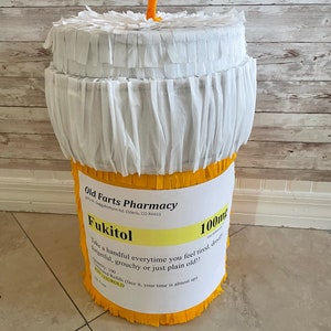 May include: A cylindrical pi&ntilde;ata shaped like a prescription pill bottle with a white fringed lid and a yellow fringed base. The label reads Old Farts Pharmacy, Fukitol 100mg, with instructions to take a handful when feeling tired, droopy, or old.