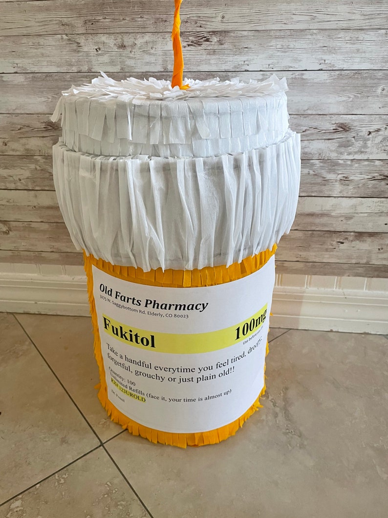 May include: A cylindrical pi&ntilde;ata shaped like a prescription pill bottle with a white fringed lid and a yellow fringed base. The label reads Old Farts Pharmacy, Fukitol 100mg, with instructions to take a handful when feeling tired, droopy, or old.