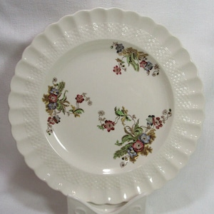 以下が含まれることがあります： ピンク、グリーン、ブルーの色合いの花柄が織り込まれた白いセラミック製のディナー皿です。