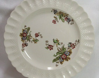 Op de afbeelding: Een wit keramisch dinerbord met een geweven patroon en bloemmotieven in tinten roze, groen en blauw.