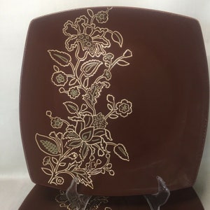 Puede incluir: Plato cuadrado marrón con un diseño floral en dorado y verde oliva. El patrón decorativo presenta flores, hojas y enredaderas estilizadas. El plato forma parte de un juego y se exhibe sobre un soporte transparente.