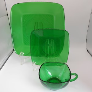 Può includere: Un set di tre piatti quadrati in vetro verde. Il piatto più grande è sullo sfondo, il piatto medio è al centro e il piatto più piccolo è in primo piano. Tutti e tre i piatti hanno un design rialzato attorno al bordo.