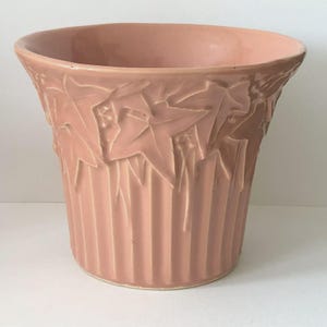 May include: A blush pink ceramic planter with a flared rim and a textured leaf design. The planter has vertical ribbed detailing on the lower portion. This decorative planter is suitable for indoor or outdoor use.