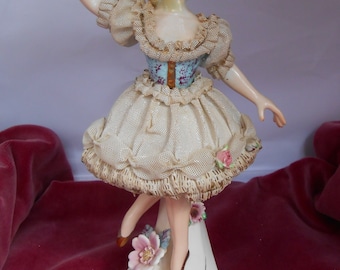 May include: A porcelain figurine of a ballerina wearing a white dress with lace trim and a blue and white bodice. She is standing on a white base with floral accents.