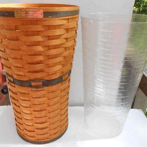 May include: A woven, light brown basket with a dark brown metal band and handle. The basket has a rectangular tag that reads "Longaberger". A clear plastic liner is next to the basket.