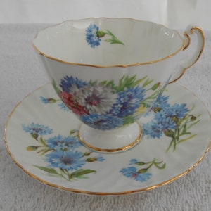 May include: A white teacup and saucer with a floral pattern of blue, pink, and white flowers. The cup and saucer have a gold trim.
