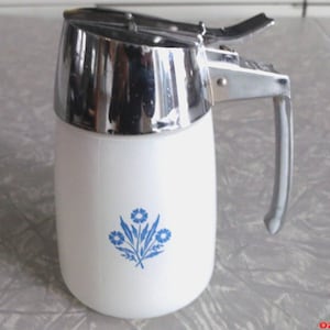Op de afbeelding: Witte siroopdispenser met een chromen deksel en handvat. De dispenser heeft een blauw bloemenontwerp op de voorkant. De handgreep is gebogen en het deksel heeft een scharnierende opening. Een klassiek keukenartikel.