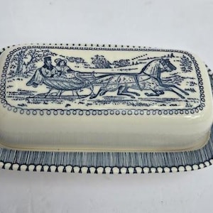 Op de afbeelding: Een rechthoekige keramische botervloot met een crèmekleurige basis en een deksel met een blauwe transferprint van een door een paard getrokken arrenslee in een winterlandschap. Het deksel en de randen van de schaal hebben een blauwe rand.