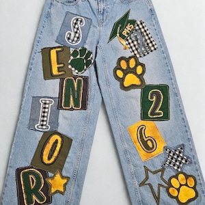 May include: Light blue denim jeans decorated with patches. Patches include the letters "SENIOR", paw prints, stars, a graduation cap, and the number "26". The patches have various colors and patterns.