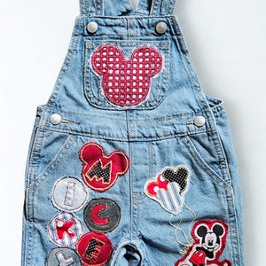 May include: Light blue denim overalls with Mickey Mouse and letter patches. The bib pocket features a red Mickey Mouse head. Patches include the letters M, I, C, K, E, Y, and a Mickey Mouse graphic.