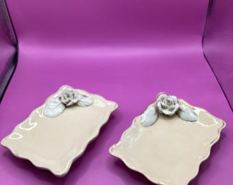 May include: Two off-white rectangular ceramic dishes with a scalloped edge and a rose design. The roses are white with light green leaves. The dishes have a gold trim and are set against a purple background.