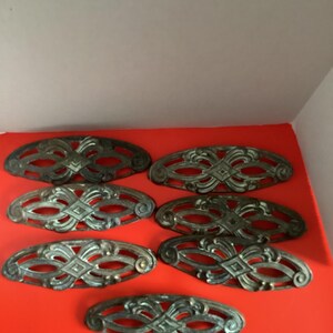 May include: A collection of seven antique bronze-toned drawer pulls. Each pull has an oval shape with an ornate, symmetrical design featuring interwoven patterns and diamond shapes. They are arranged on a bright red surface.