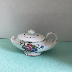 May include: A white ceramic genie lamp with a lid and handle, adorned with a floral pattern in pink, blue, and green. The lamp has gold trim and a small knob on the lid. A decorative piece.