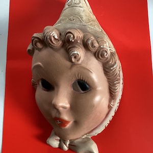 May include: A vintage ceramic wall mask of a stylized face with a bonnet and bow. The mask features a light skin tone, black eyes, red lips, and brown curly hair. The bonnet is off-white with a decorative pattern. The background is a solid red.