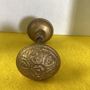 May include: A vintage brass doorknob with an ornate, textured design on the main knob. The knob has a circular shape and a warm, antique gold color. The stem and smaller knob are also brass.