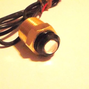 May include: A brass-colored threaded cylindrical component featuring a black circular cap with a white center lens. Attached to the rear of the metal housing are two thin black electrical wires, one of which has a small red accent near the base.