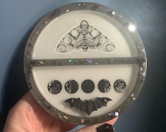 May include: A round, gray resin tray with a black bat and five black moon phases. A black and white moth with a skull design is on the top half of the tray.