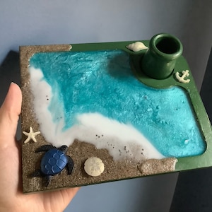 May include: A green wooden tray with a blue and white resin ocean scene. The tray features a small turtle, a seashell, a starfish, and an anchor. There is a green ceramic cup on the tray.