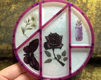 May include: A round, white, resin tray with six compartments. The tray has a purple glitter rim and features a black butterfly, a black rose, a bottle of potion, and a bouquet of flowers.