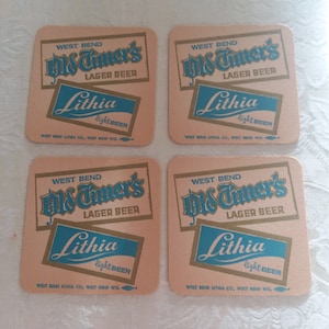 May include: Four square coasters with a light pink background. Each coaster features a gold and blue logo with the text "Old Timer's Lager Beer" and "Lithia Light Beer". The text "West Bend Lithia Co. West Bend Wis." is printed on the bottom of each coaster.