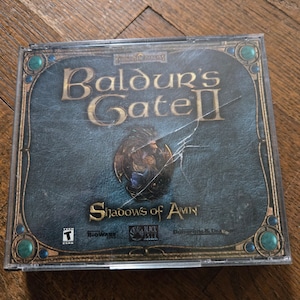 May include: A CD case for the video game Baldur's Gate II: Shadows of Amn. The case is made of clear plastic and has a blue and gold design. The game is rated T for Teen by the ESRB. The game is published by BioWare and distributed by Black Isle Studios.