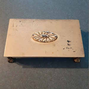 May include: A white metal trinket box with a decorative oval design on the lid and four small feet. The box has a worn, vintage look.