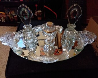 May include: A vintage crystal vanity set with three bottles and a mirrored tray. The bottles have ornate stoppers and are decorated with cut glass. The tray has a scalloped edge and is decorated with a floral design.