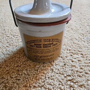 May include: A white ceramic crock with a wire handle and a brown label that reads "Wisconsin Homestead Sharp Cheddar Cold Pack Cheese Food". The label also includes the ingredients, distributor, and net weight of 20 ounces (1 lb. 4 oz.).