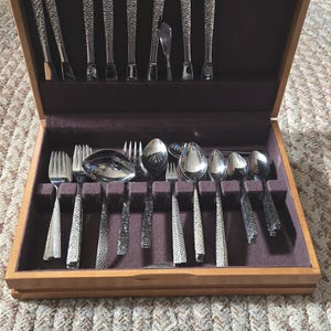 May include: A wooden silverware chest with a dark purple velvet interior, holding a complete set of stainless steel flatware. The flatware features textured handles and includes forks, spoons, and knives. The chest is open, revealing the neatly arranged utensils.