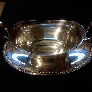 May include: Silver gravy boat with a matching saucer. The boat has two handles and a smooth, polished surface. The saucer has a decorative edge.