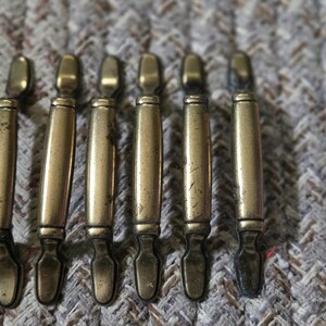 May include: A set of six brass-toned metal drawer pulls arranged in a vertical row. Each pull features a slender, cylindrical central bar with a smooth finish, flanked by decorative, flared ends that taper to a point at the top and bottom.