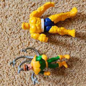 May include: Two yellow and green plastic action figures of superheroes. The first figure is a yellow humanoid with a blue and white belt. The second figure is green with yellow accents and has multiple gray tentacles.