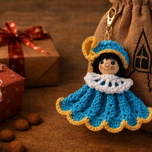 Könnte beinhalten: Eine Häkelpuppe als Schlüsselanhänger mit schwarzem Haar, einer blauen Mütze mit gelbem Rand, einem weißen Spitzenkragen und einem blauen Kleid mit gelbem Wellensaum. Die Figur ist an einem goldfarbenen Metallkarabiner befestigt.