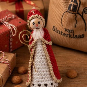 Op de afbeelding: Een gehaakt Sinterklaasfiguur met een rood en wit gewaad, een goudkleurige mijter en een witte baard. Het figuur houdt een goudkleurige metalen staf vast. Daarnaast staat een beige stoffen zak met de tekst "van Sinterklaas".