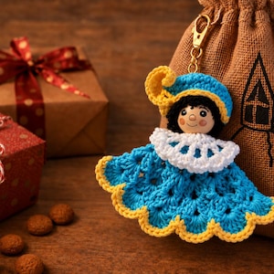 Puede incluir: Un llavero de muñeca de ganchillo con cabeza de madera, pelo negro y gorro azul con pluma amarilla. La muñeca lleva un cuello blanco con volantes y un vestido azul con borde festoneado amarillo, unido a un cierre de metal dorado.