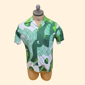 May include: A short-sleeved button-down shirt featuring a bold, abstract pattern in shades of forest green, lime green, and white. The shirt has a classic collar, a single chest pocket on the left side, and white buttons running down the front center.
