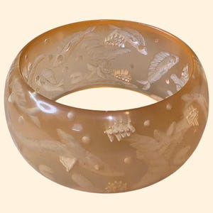 May include: A translucent, wide bangle bracelet in a warm, amber hue. The bracelet features an intricate design of etched fish and aquatic plants. The details are highlighted with a lighter, opaque color, creating a contrast against the amber background.